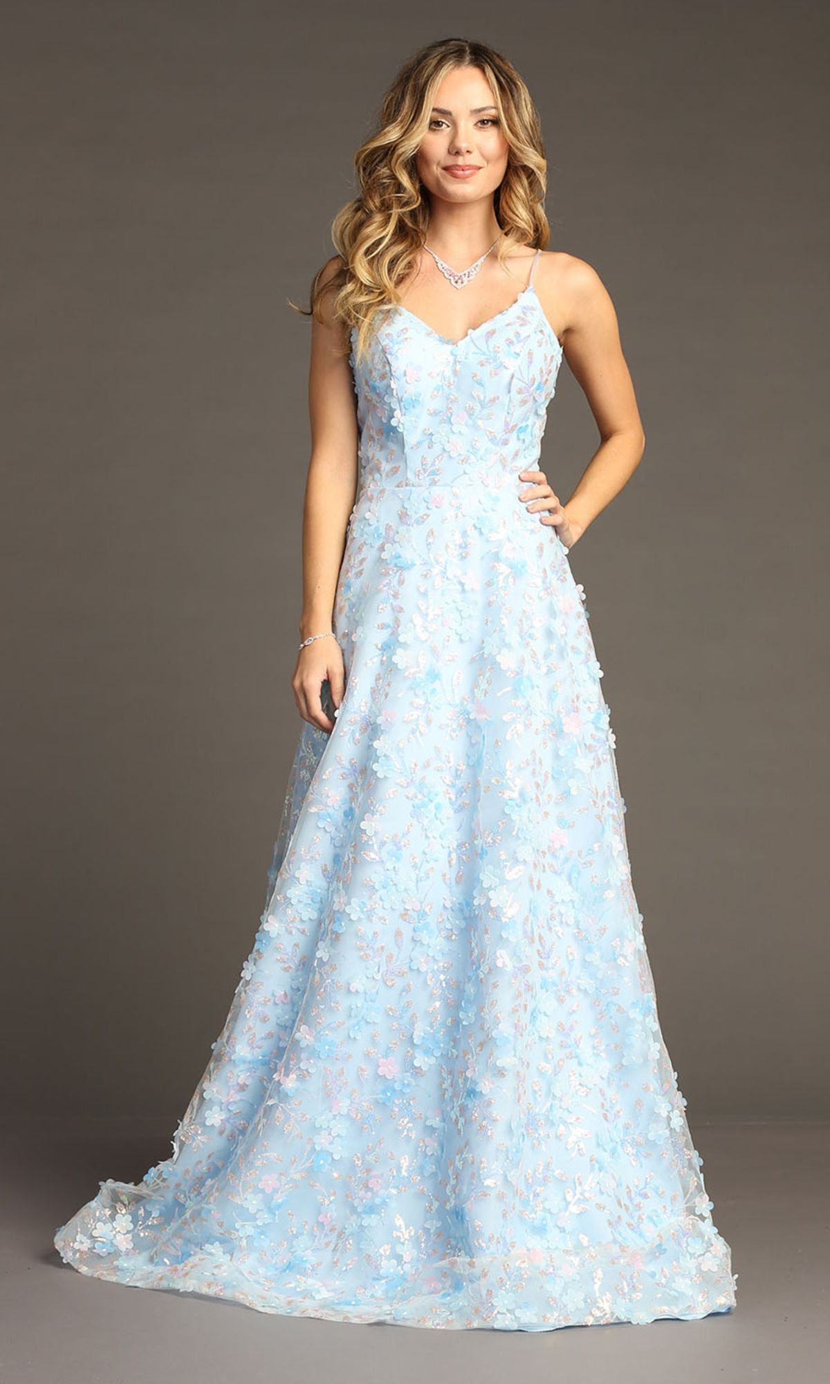 A woman wears the Chicas CHF3092 long light blue floral prom dress, featuring delicate 3-D flowers, thin straps, and a v-neckline, standing against a plain gray background.